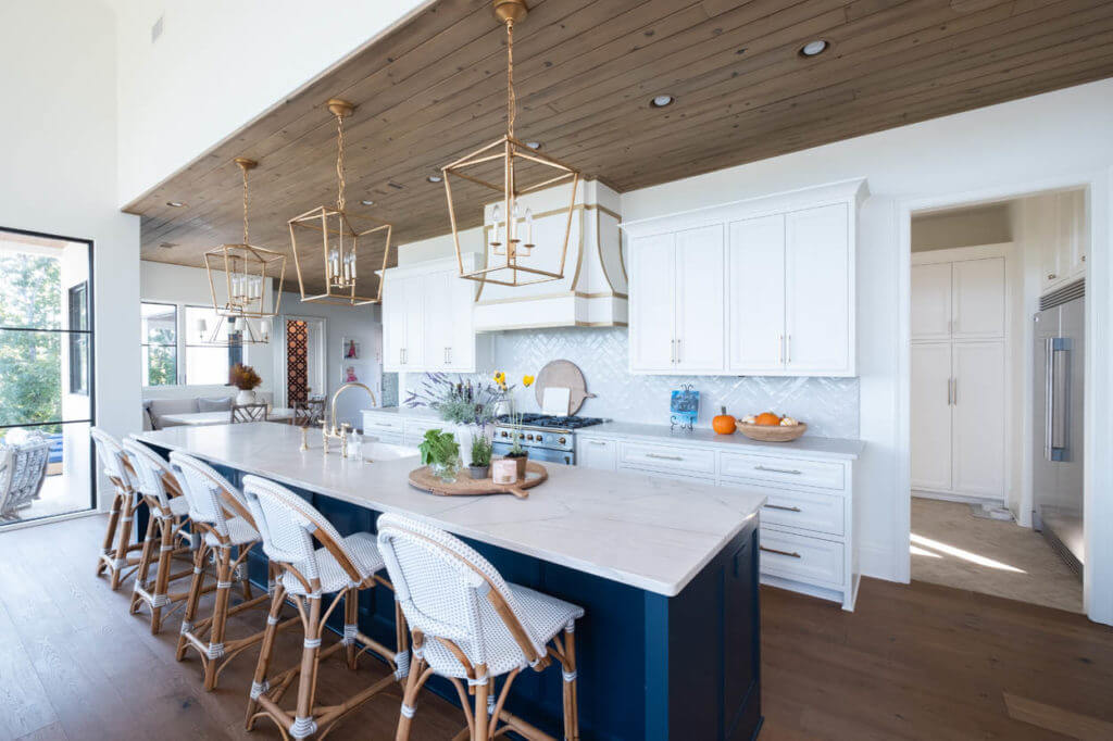 Open kitchen with gold hanging lights, contrasting, curvy vent hood, and tongue and groove dark brown ceiling.